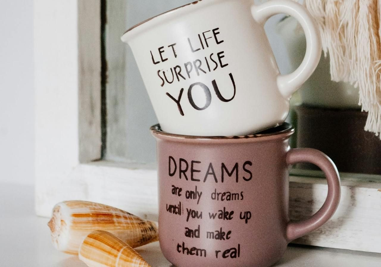 Two coffee mugs displaying the phrases "Let life surprise you" and "Dreams" in a cozy setting.